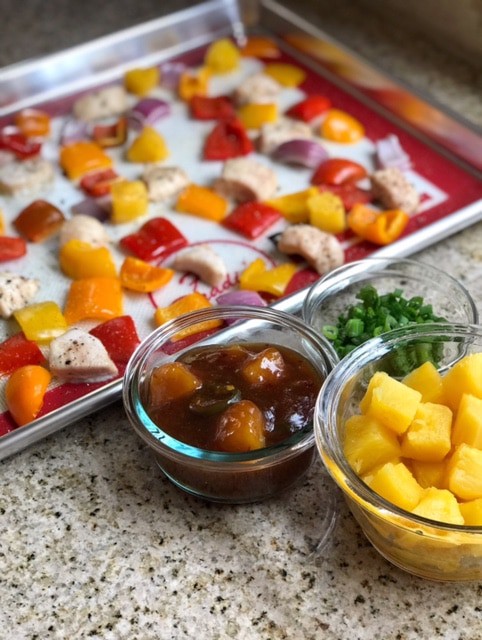 Side-view of sheet pan pineapple chicken next to bowls of sauce, scallions, and pineapple pieces