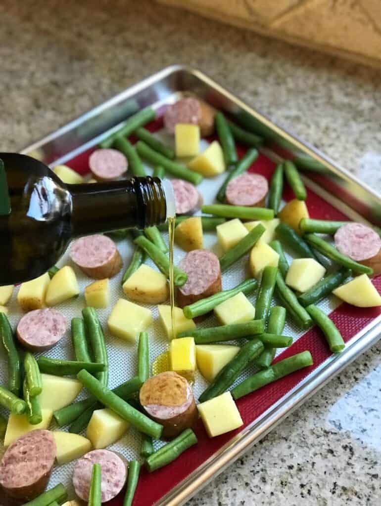 Close-up shot of oil being drizzled over a sheet pan with sausages, potatoes, and green beans